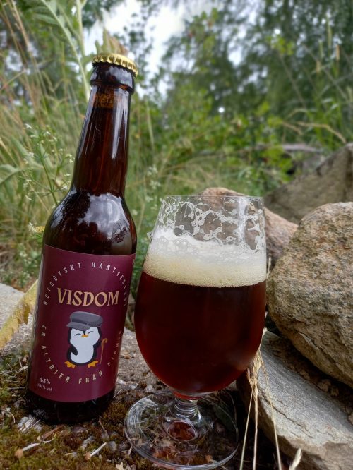 Ett glas Visdom Schwarsbier står bredvid en flaska visdom. Bakgrunden är lummig.